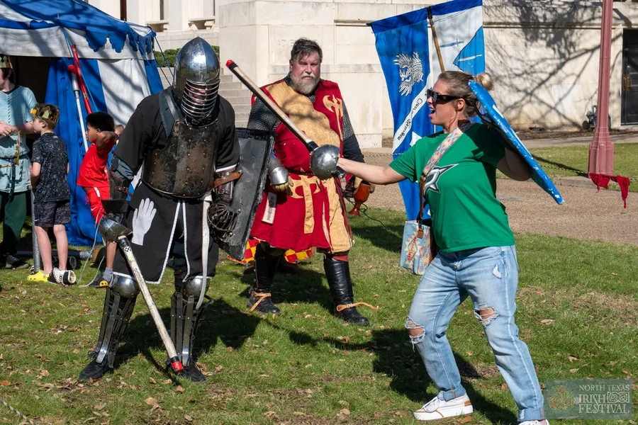 A festival attendee play-fighting with a costumed knight in full armor on the fairgrounds lawn.