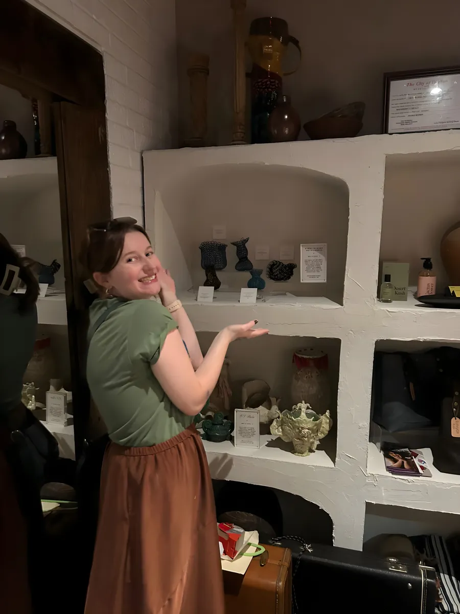 Local artist Avery McGhee smiling and gesturing toward a display of her metalwork on shelves.